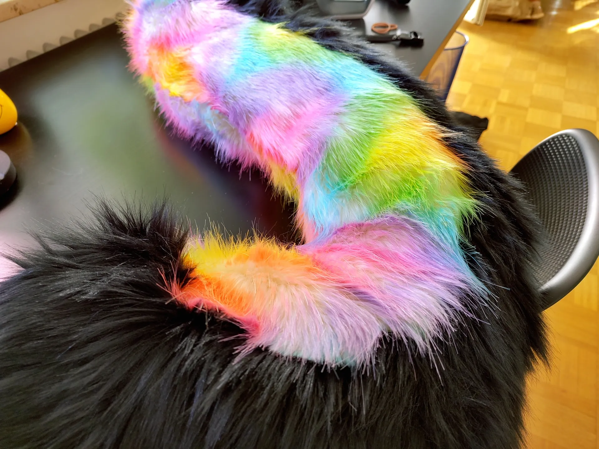 Close up of a fursuit tail, the bottom is black while the top is rainbow colored. It's lying on my way too clean desk.